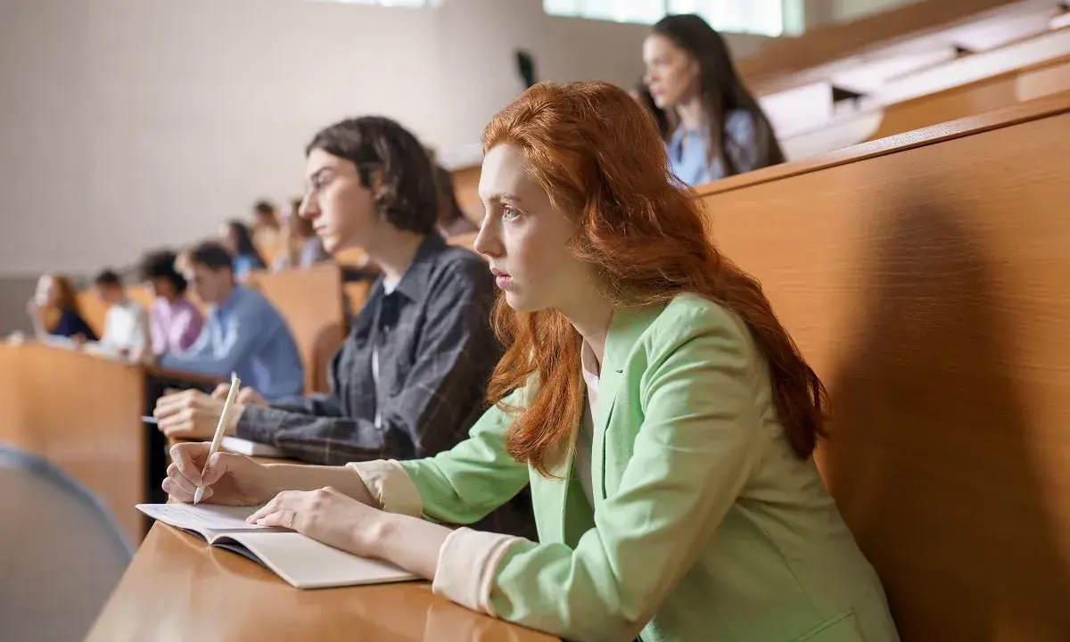 Studentka z rudymi włosami skupiona na wykładzie, robi notatki. Jej kolega obok również studiuje psychologię.