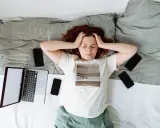 Kobieta z rękami na skroniach leży na łóżku otoczona telefonami i laptopem. Widać wyraźny stres przed pracą.