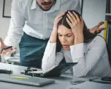 Kobieta z rękami na głowie, przytłoczona stresem w pracy. Jej szef gestykuluje, co potęguje jej cierpienie. To moment, gdy rezygnacja z pracy z powodu stresu wydaje się jedynym wyjściem.