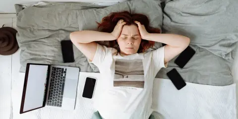Kobieta z rękami na skroniach leży na łóżku otoczona telefonami i laptopem. Widać wyraźny stres przed pracą.