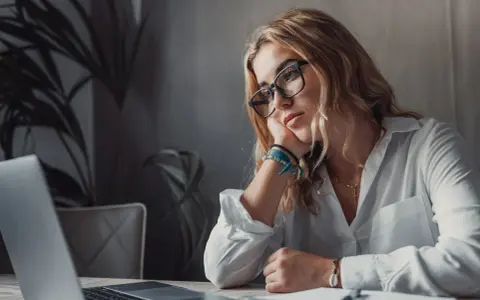Zamyślona kobieta przy laptopie, z podpartą głową na dłoni. Wokół niej panuje atmosfera rezygnacji, jakby mówiła sobie: "nie mam marzeń".