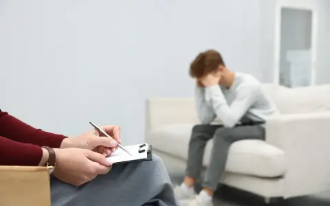 Młody mężczyzna siedzi na kanapie, ukrywając twarz w dłoniach, przeżywając cierpienie psychiczne. Obok niego terapeuta notuje coś na kartce.