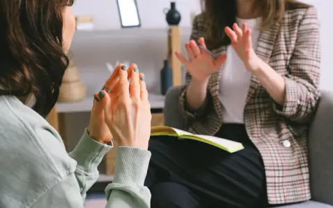 Kobieta z zielonymi paznokciami gestykuluje, jakby stresowała się byle czym. Obok niej siedzi druga osoba w kratkę.