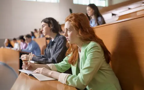 Studentka z rudymi włosami skupiona na wykładzie, robi notatki. Jej kolega obok również studiuje psychologię.