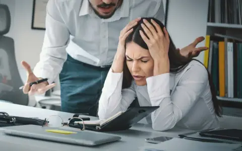 Kobieta z rękami na głowie, przytłoczona stresem w pracy. Jej szef gestykuluje, co potęguje jej cierpienie. To moment, gdy rezygnacja z pracy z powodu stresu wydaje się jedynym wyjściem.