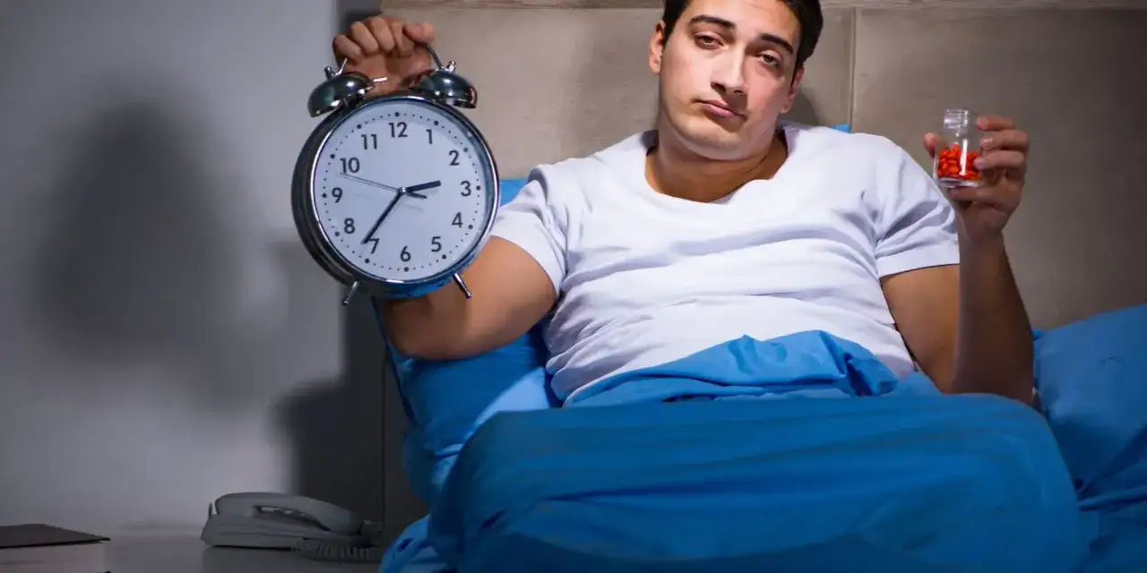 Zmęczony mężczyzna w łóżku trzyma budzik i tabletki, co może sugerować zaburzenie adaptacyjne i problemy ze snem.